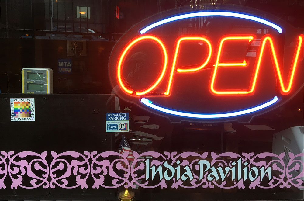 The 100% sign is seen in the window of the India Pavillion restaurant beside a large neon open sign and an ornate pink border with the name of the restaurant