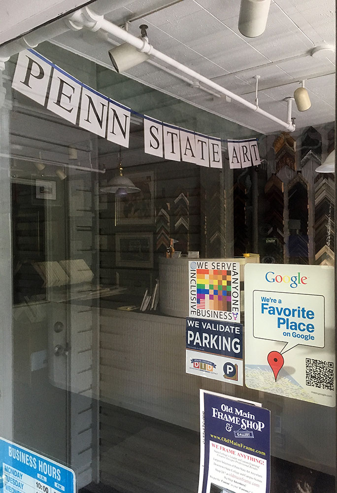 The 100% Sign is seen on the entry door to the Old Main Frame shop. Through the door a banner advertises Penn State Art and we see picture frame samples on the wall