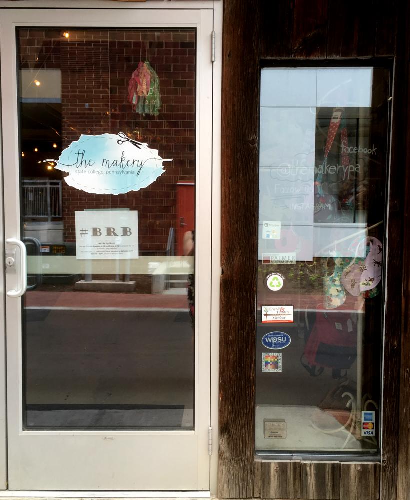 The 100% sign is seen in a front window adjacent to the entry door of The Makery